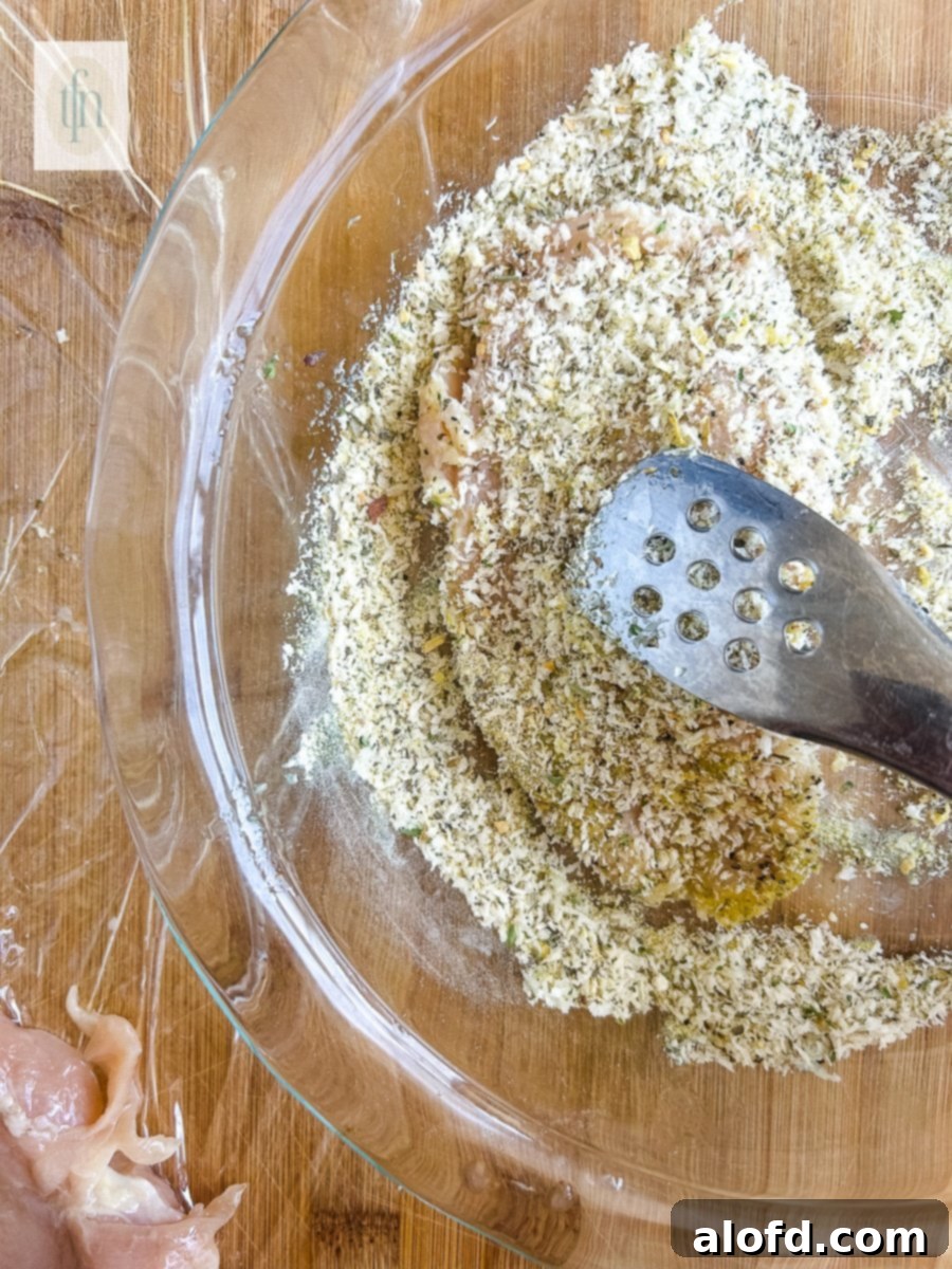Golden Panko-Crusted Chicken Breast 5 A hand pressing a chicken breast into panko breading in a shallow dish.