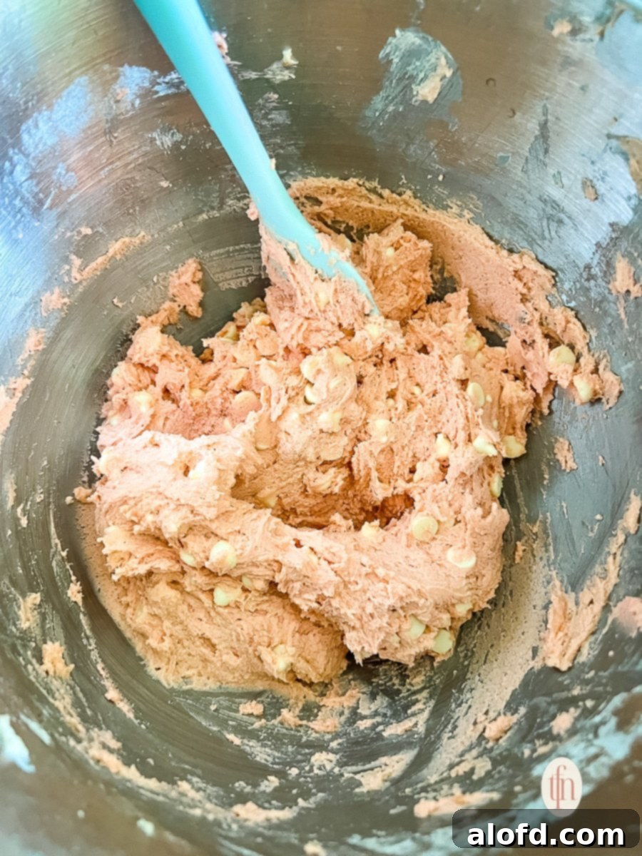 Strawberry Cheesecake Cookie Bliss 6 Blue mixing spoon stirring white chocolate chips into bowl of strawberry cookie dough, highlighting the vibrant pink color.