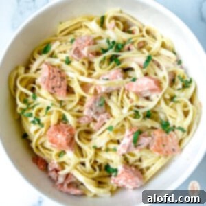 Decadent Salmon Alfredo 11 Flaky bite-sized pieces of baked salmon on top of creamy fettuccine in a white bowl, ready to be enjoyed.