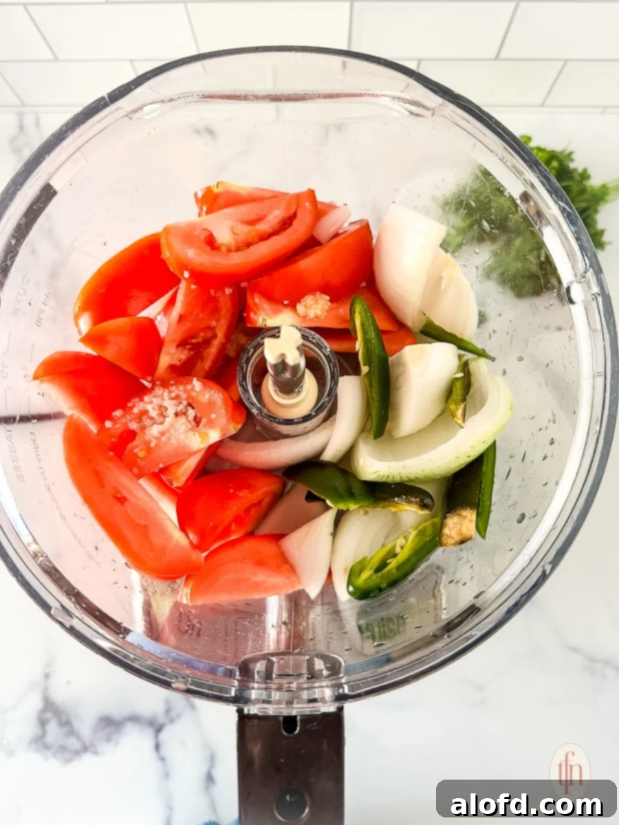 Chunks of perfectly processed tomatoes, white onion, and green chiles in the bowl of a food processor, showing the ideal texture before final blending.