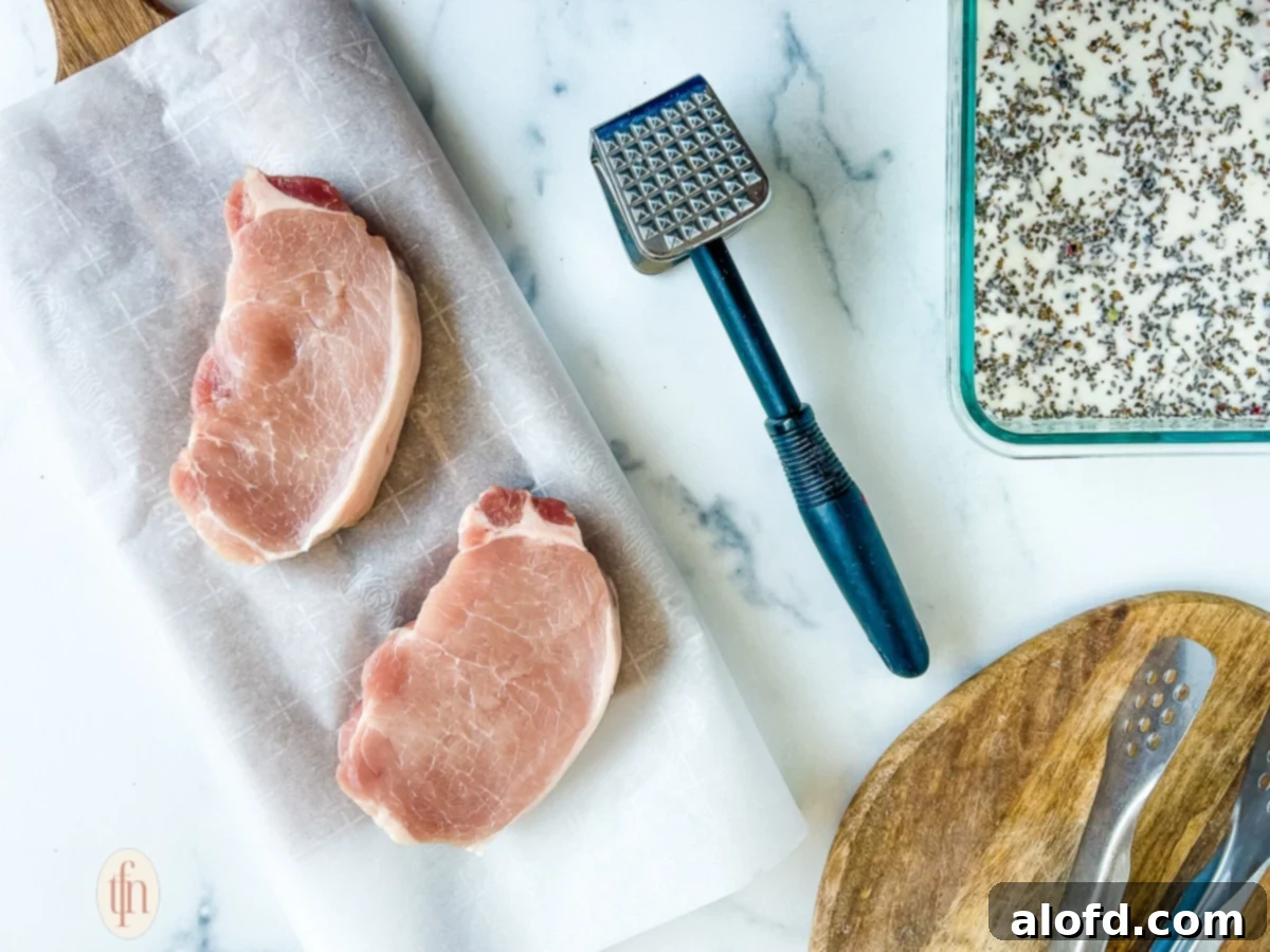 Two raw, thick-cut pork chops resting on a wooden cutting board, positioned next to a stainless steel meat mallet.