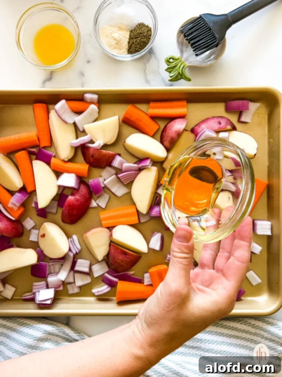 A hand expertly drizzling cooking oil over a colorful mix of chopped vegetables spread on a sheet pan, ensuring even coating.