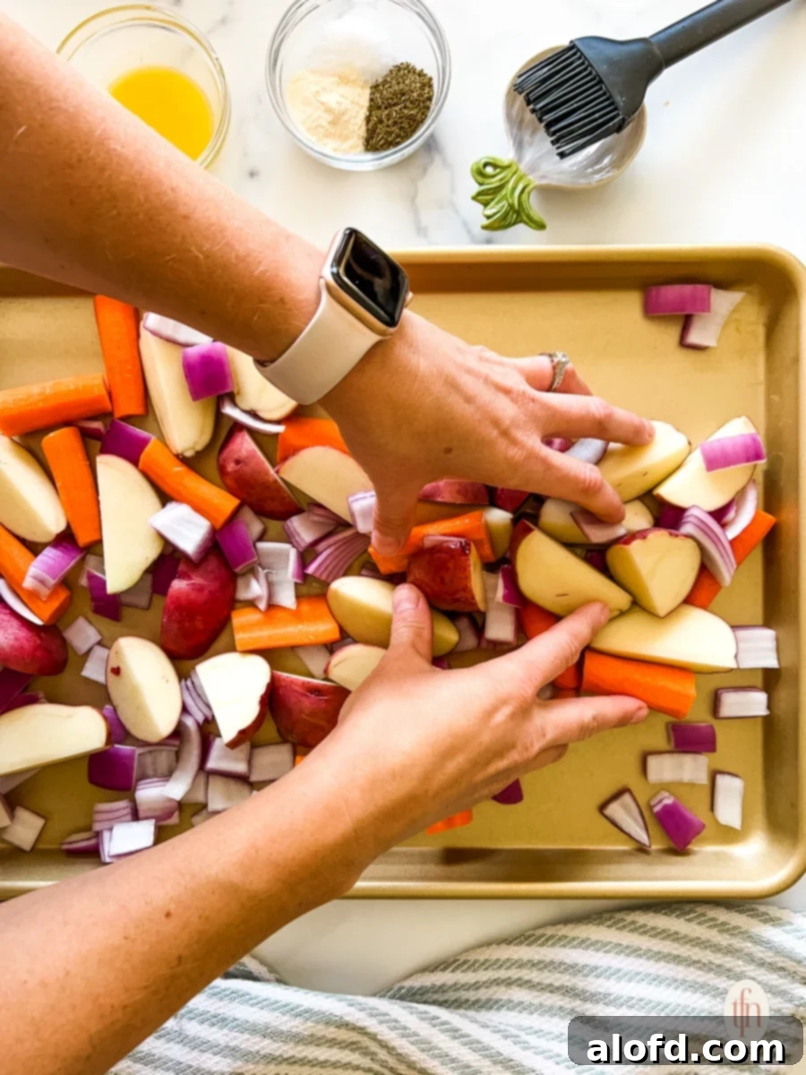 Hands carefully arranging seasoned chicken thighs and colorful chopped vegetables onto a single sheet pan, ready for the oven.