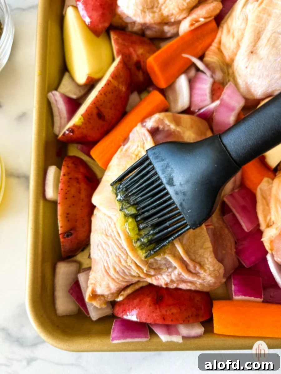 A pastry brush applying melted butter to the surface of a raw chicken thigh on a baking sheet, preparing it for roasting.