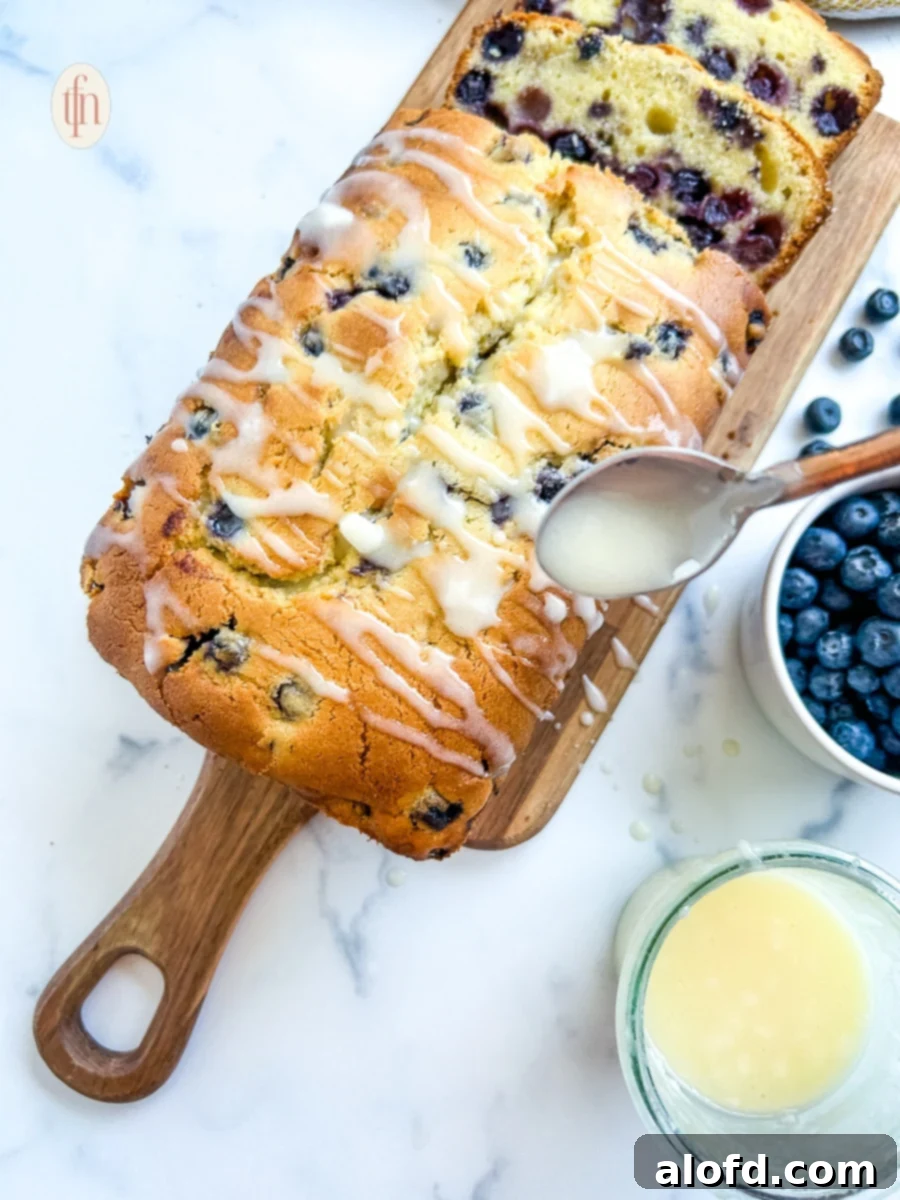 Luscious Blueberry Pound Cake 2 Using a spoon to drizzle glaze over a loaf cake with blueberries.