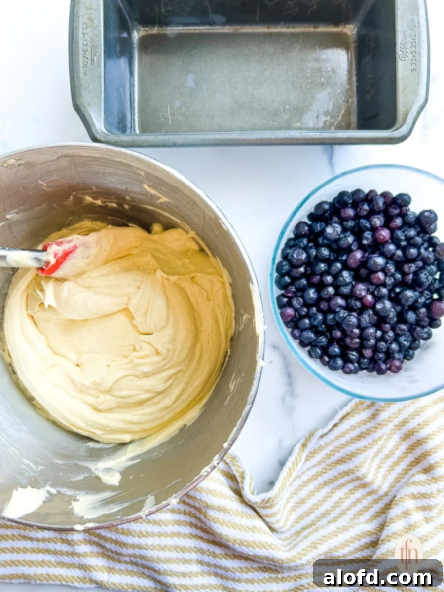 Luscious Blueberry Pound Cake 7 Overhead shot of an empty loaf pan, bowl of frozen blueberries, and a stand mixer bowl with pale yellow batter in it.
