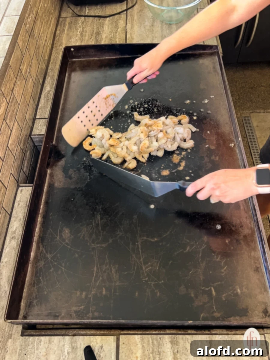 Raw shrimp being skillfully flipped with two metal spatulas on a hot Blackstone griddle.