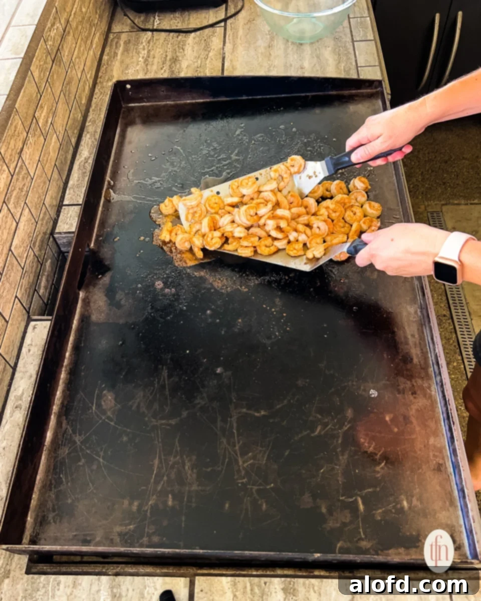 Cooked shrimp, now pink and opaque, being stirred and flipped with metal spatulas on the griddle, ready to be removed.