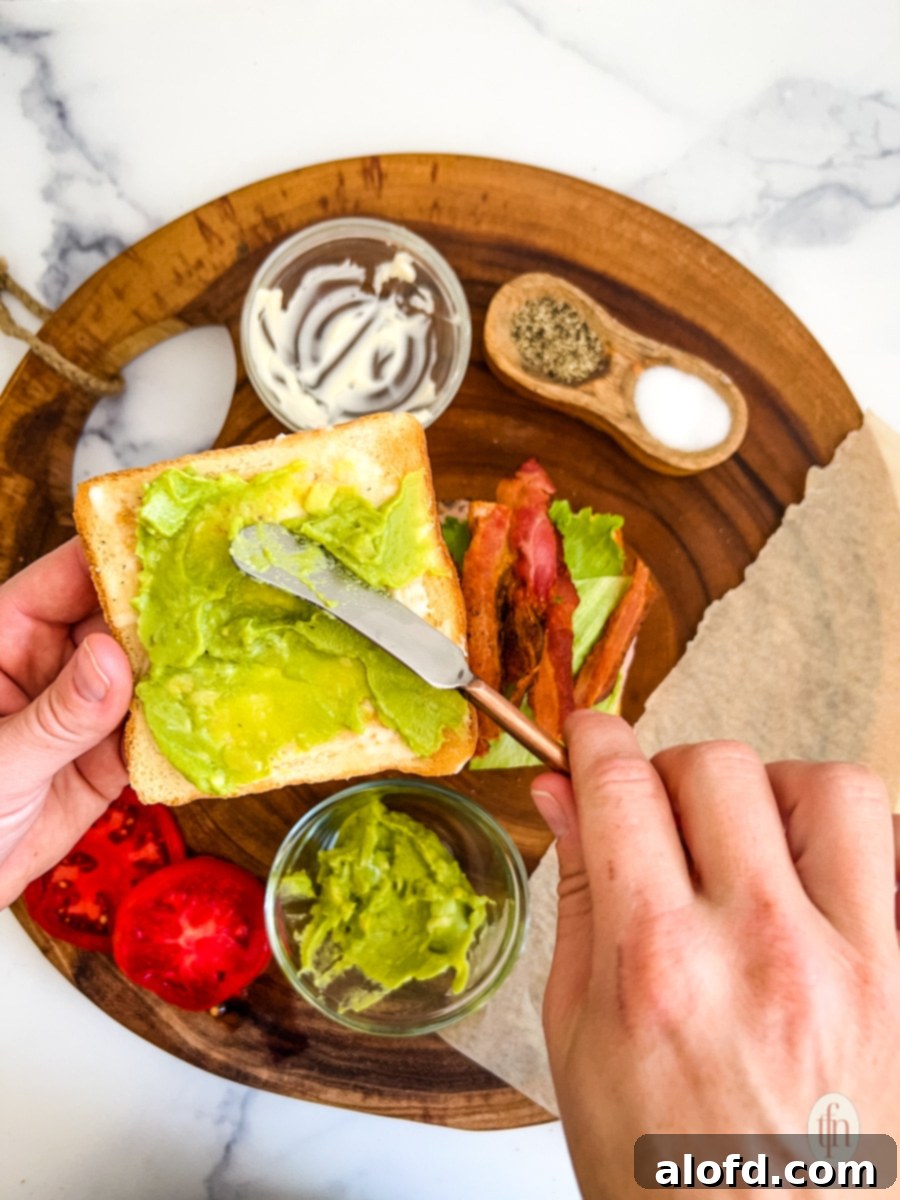 Spreading sauce on bread while assembling the ingredients for a chicken club sandwich on a round wooden board.