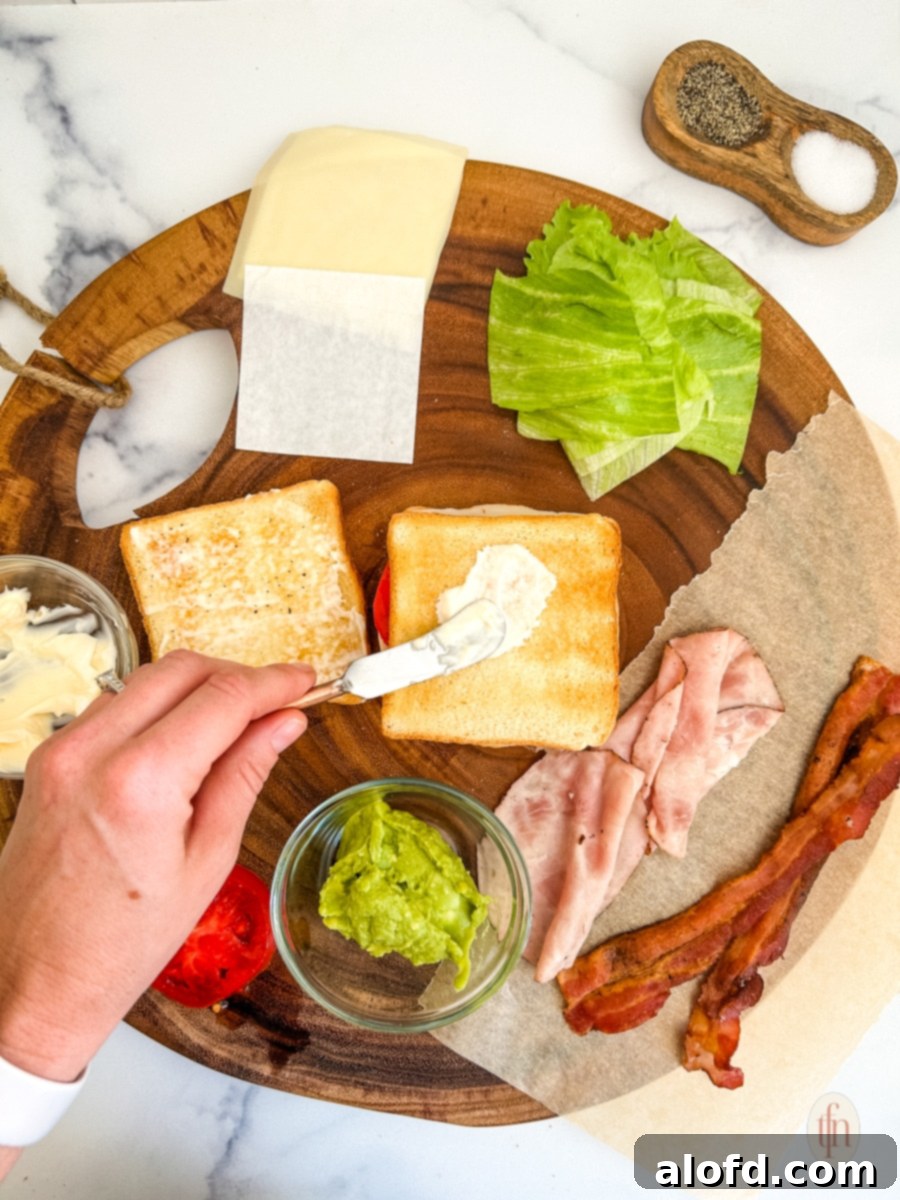 Spreading sauce on bread with prepared ingredients for chicken club sandwich on a round wood board.