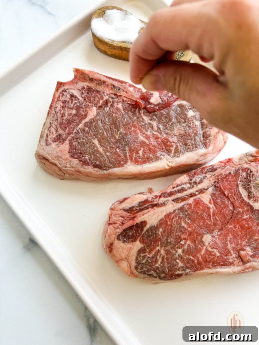 A cook generously seasoning a raw strip steak with coarse salt and black pepper just before cooking, ensuring even coverage.