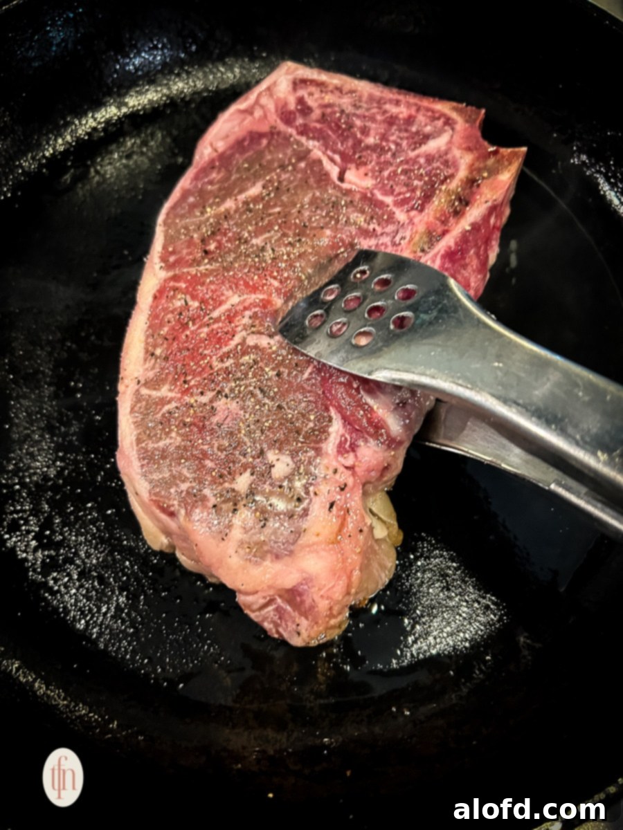 A chef using tongs to carefully turn over a bone-in NY strip steak as it cooks in a cast iron skillet, showcasing a perfect golden-brown sear.