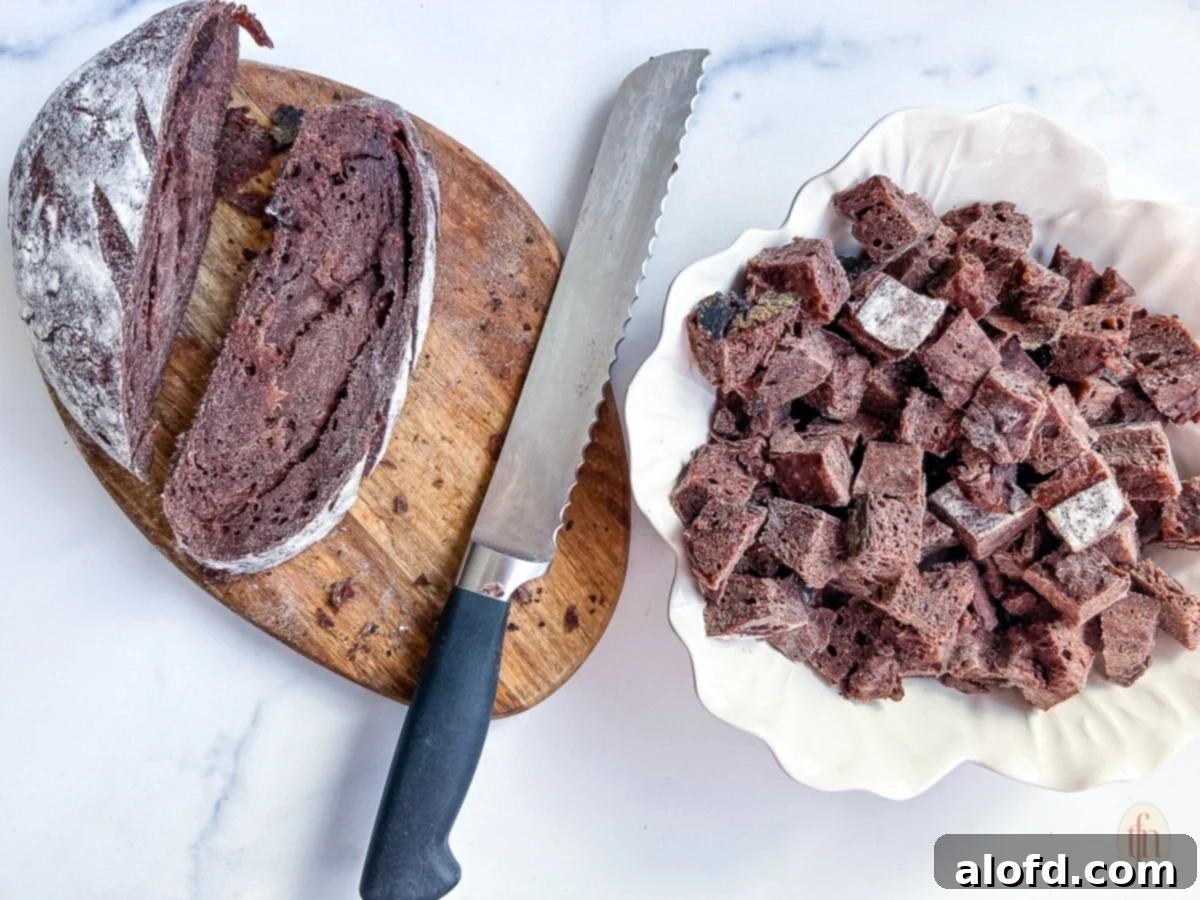 Slicing chocolate sourdough bread into cubes with a long serrated knife on a cutting board, preparing for bread pudding.