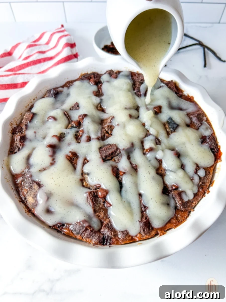 Drizzling vanilla cream sauce over chocolate sourdough bread pudding in a white pie dish, ready for serving.