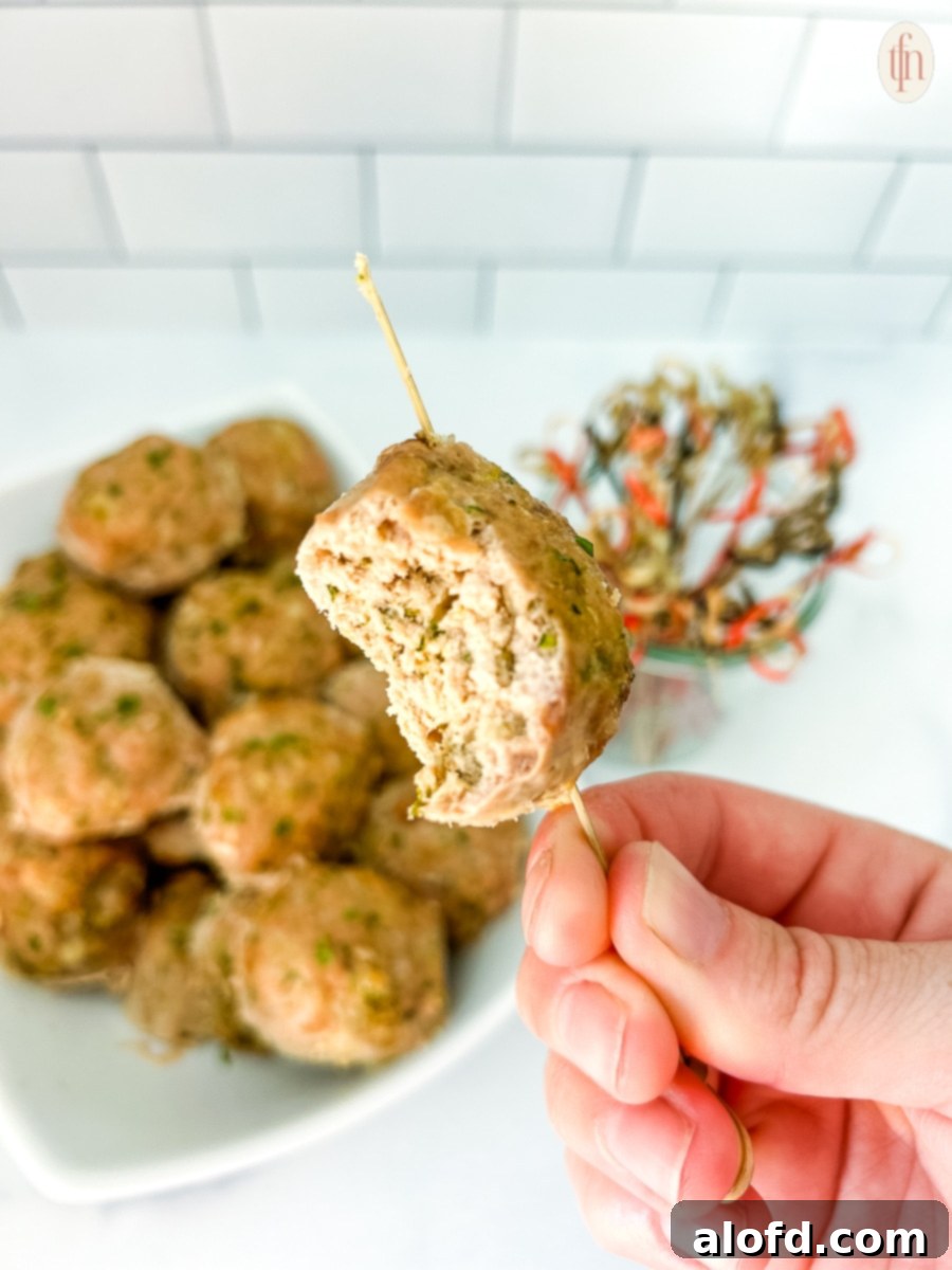 A hand holding up a turkey zucchini meatball on a toothpick with a bite taken out, showcasing its tender interior.