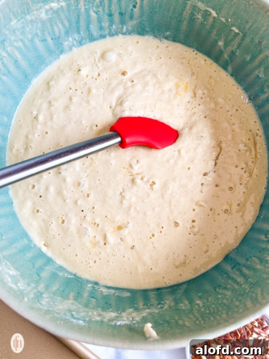 Effortless Sheet Pan Pancakes from Mix 5 A blue mixing bowl containing pancake batter, being gently whisked with a red silicone spatula, showcasing the consistency of a well-mixed batter with a few small lumps.
