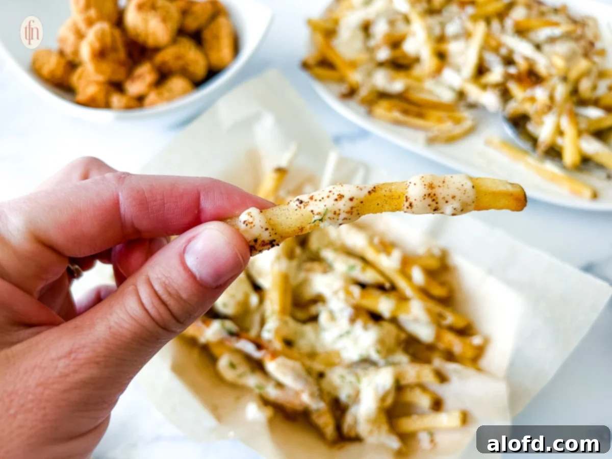 Voodoo Kitchen Fries 8 A hand holding up a single French fry, perfectly drizzled with white Cajun ranch sauce, above a plate piled high with more Voodoo Fries.