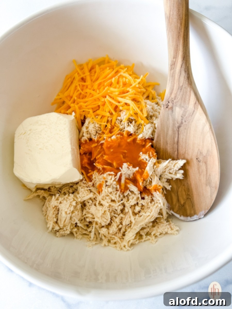 Fiery Buffalo Chicken Poppers 6 A white bowl containing a creamy mixture of shredded chicken, softened cream cheese, shredded cheddar cheese, and buffalo sauce, with a wooden spoon partially submerged, ready for filling the biscuit dough.