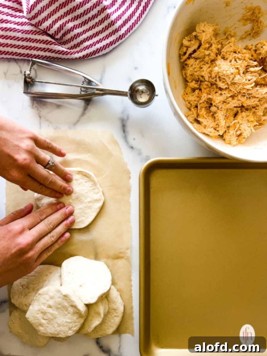 Fiery Buffalo Chicken Poppers 7 A hand using a rolling pin to flatten biscuit dough on a lightly floured kitchen counter, preparing it for filling.