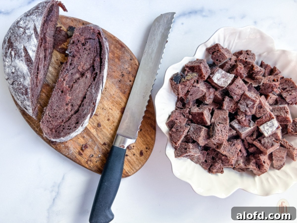Velvety Vanilla Cloud 6 Slicing chocolate sourdough bread into cubes with a long serrated knife on a cutting board.