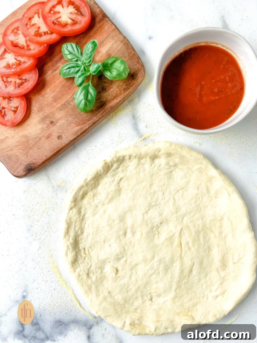 Sourdough discard pizza dough no yeast on a floured countertop next to a bowl with pizza sauce, tomato slices, and fresh basil leaves on a cutting board.