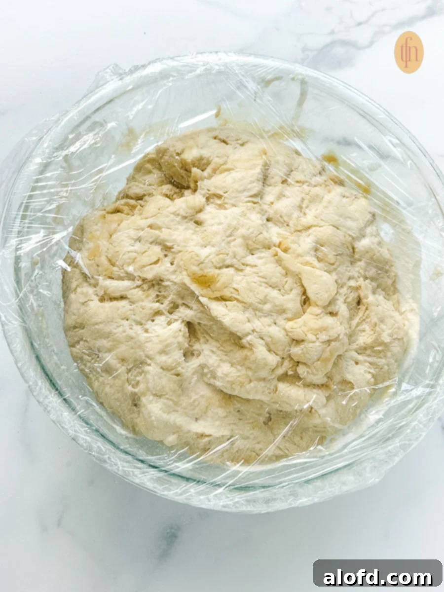 Bowl of Sourdough Discard Pizza Dough rising in a glass bowl covered with plastic wrap.