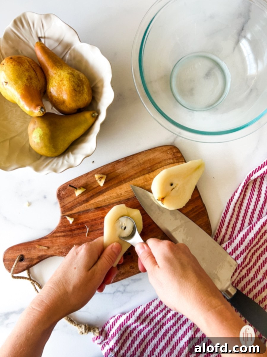 Perfect Pear Slices Every Time 7 A hand scooping out the seeds and core from a halved pear using a small spoon, with another pear half visible nearby.