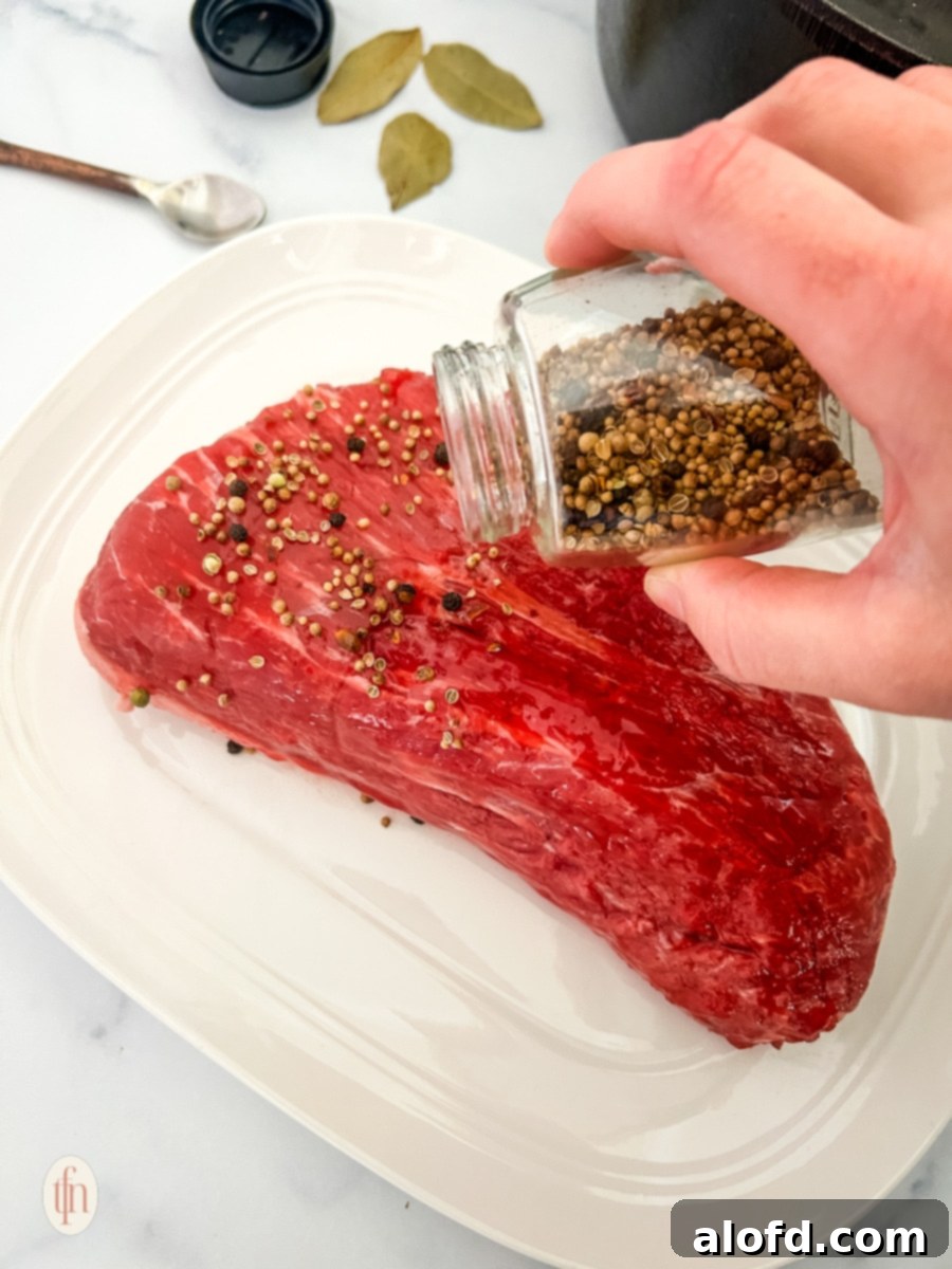Slow-Braised Dutch Oven Corned Beef 7 Seasoning being poured on top of a raw beef placed on a white plate.