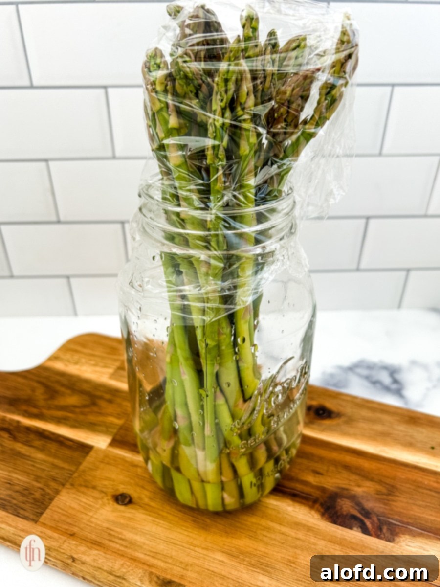 Creamy Asparagus Cheese Bake 5 Fresh asparagus spears stored upright in a glass jar with water, covered with plastic wrap, demonstrating a method for freshness.