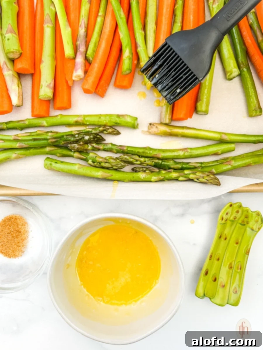 Golden Roasted Carrots and Asparagus 6 A pastry brush applying melted butter and garlic to fresh carrots and asparagus laid out on a baking sheet, preparing them for roasting.