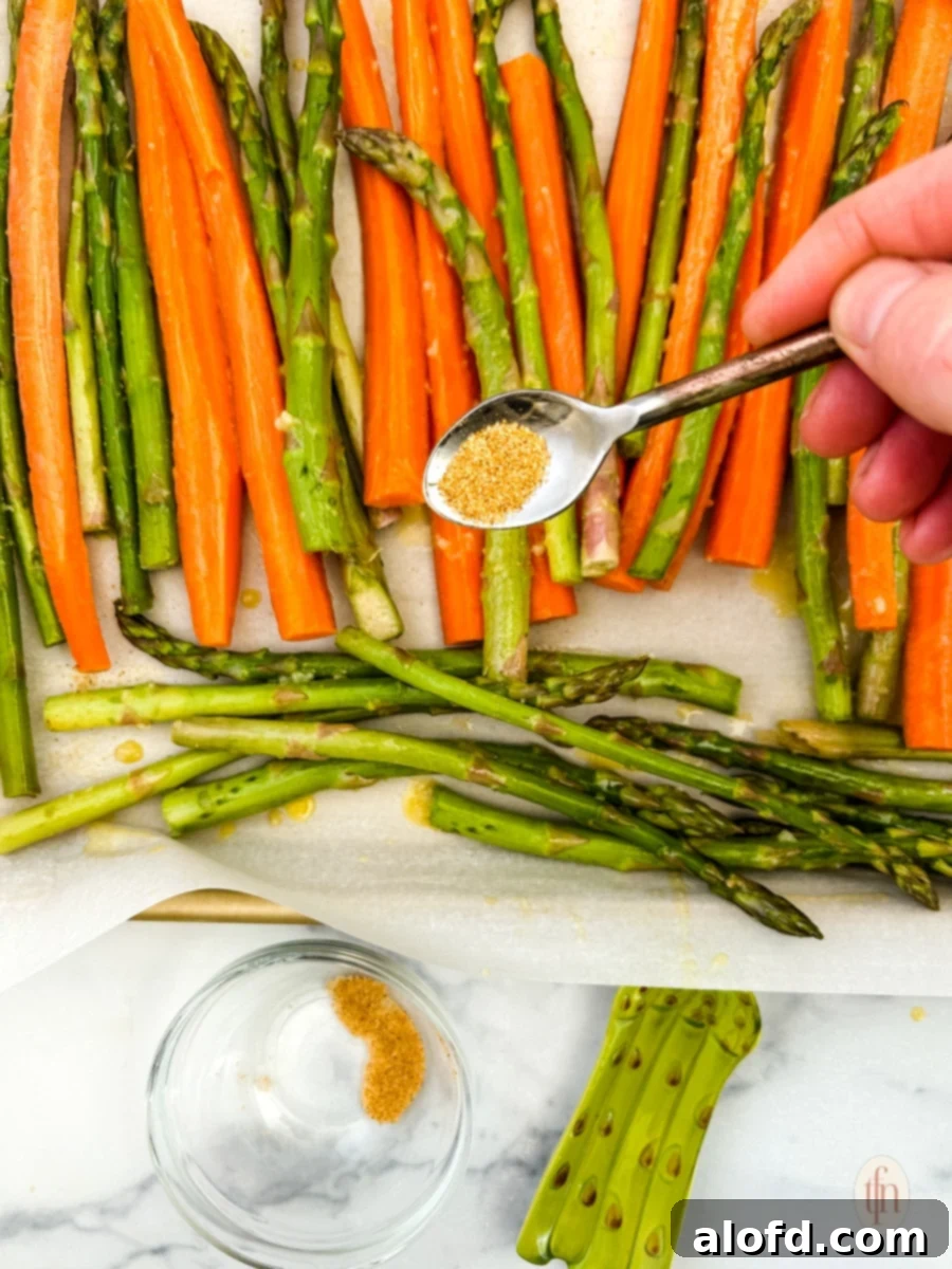 Golden Roasted Carrots and Asparagus 7 A hand sprinkling seasoning over raw carrots and asparagus spread out on a baking sheet, illustrating the final seasoning step before roasting.
