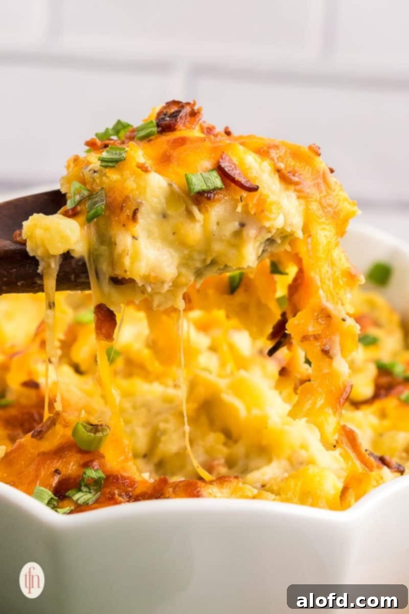 A close-up of a wooden spoon scooping a generous portion of cheesy, bacon-loaded Twice Baked Mashed Potatoes from a casserole dish, garnished with green onions.