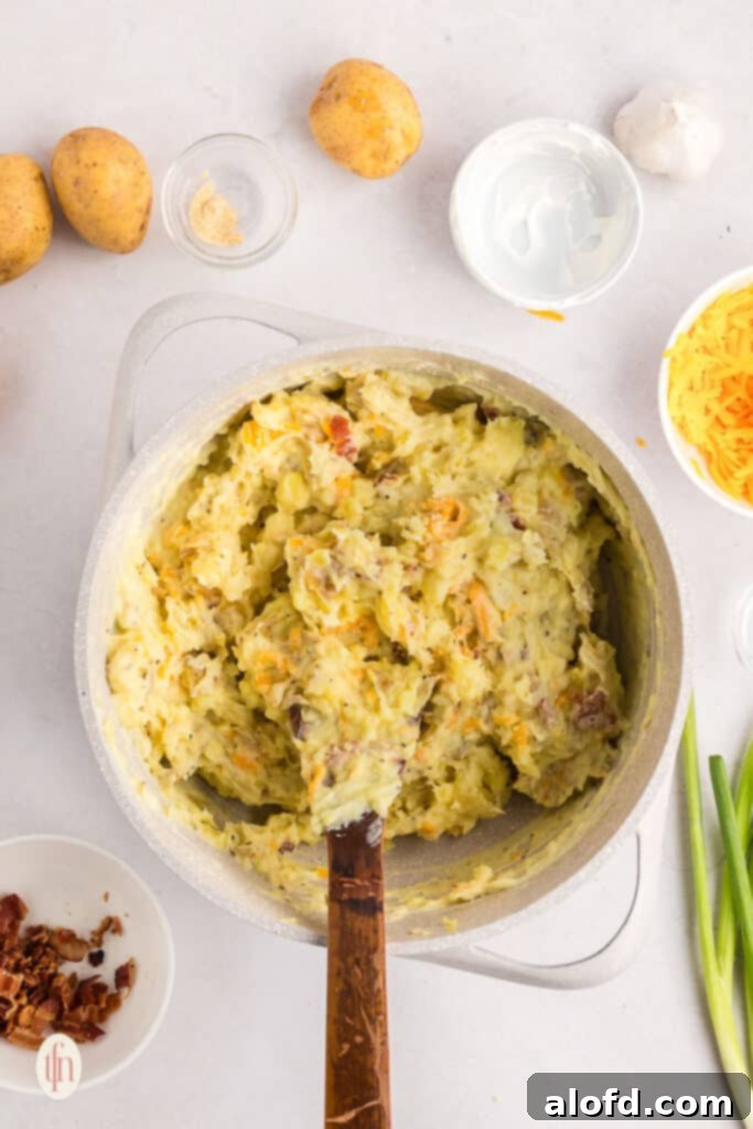 Mashed potatoes mixed with sour cream, seasonings, cheese, and bacon in a pot, before transferring to a baking dish.