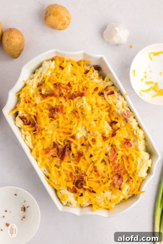 A white casserole dish filled with mashed potatoes, generously topped with shredded cheddar cheese and crispy bacon bits.