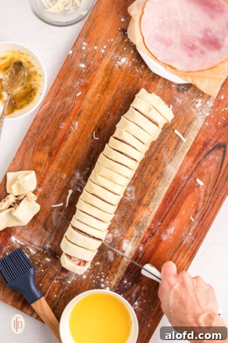 A rolled puff pastry log being sliced into individual pinwheels on a chopping board with a serrated knife.