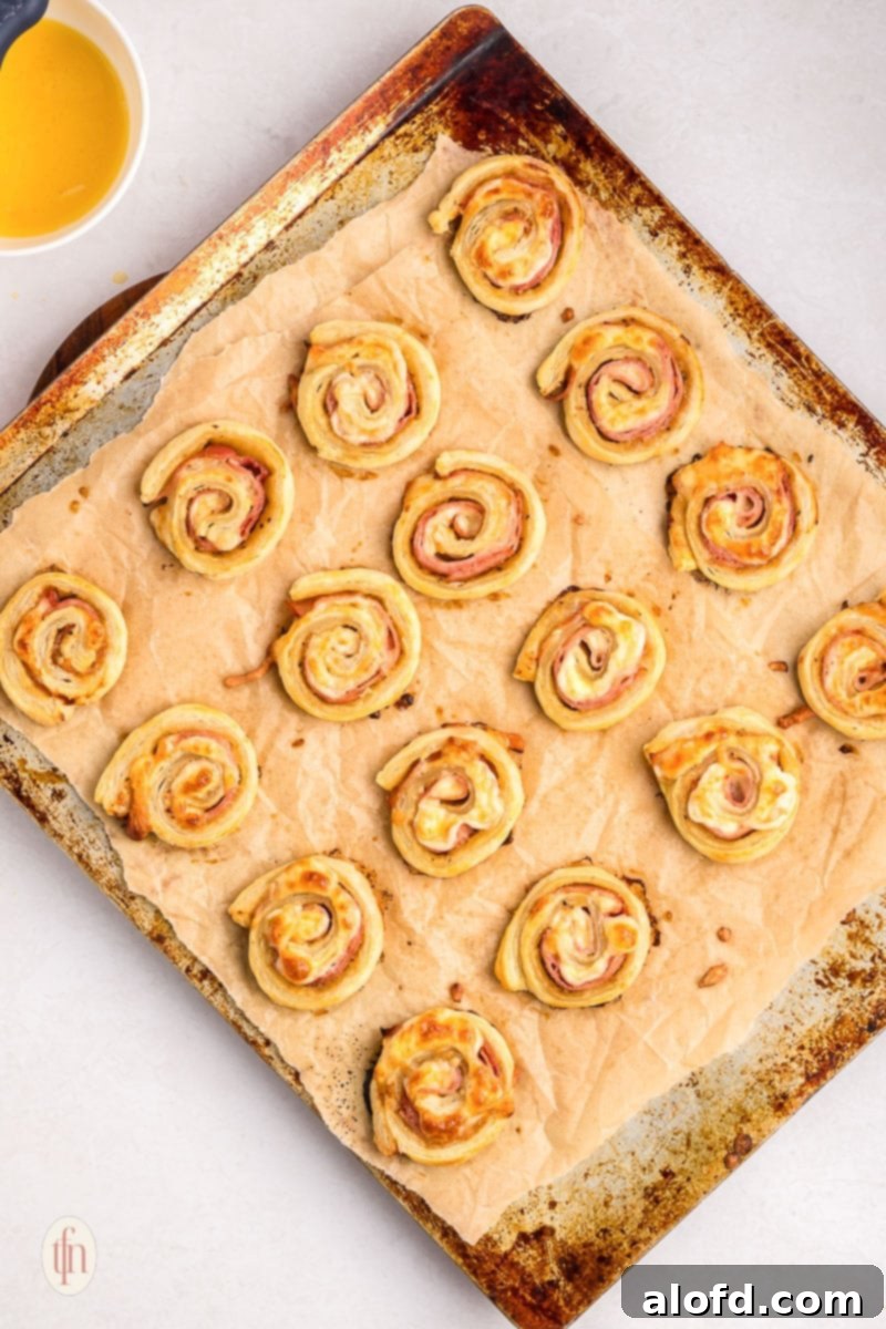 Freshly baked hot ham and cheese pinwheels, golden brown and cheesy, arranged on a baking sheet lined with parchment paper.