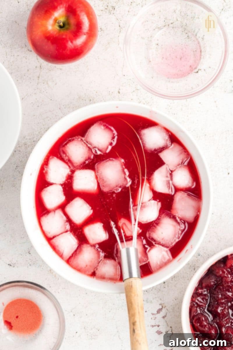 A bowl of red liquid and ice with a whisk, showing the gelatin dissolving and starting to thicken with ice cubes.
