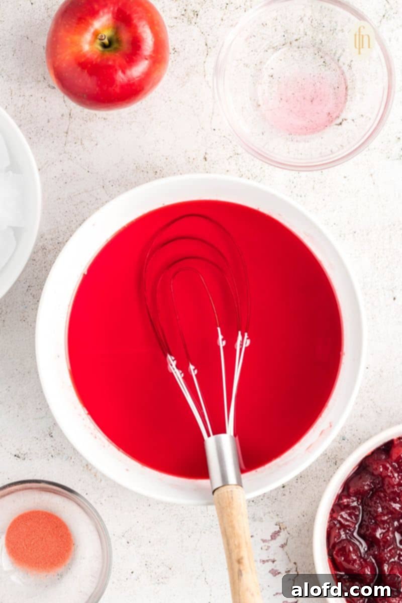 A bowl of red liquid with a whisk, showing the gelatin mixture after ice cubes have been stirred in and removed.
