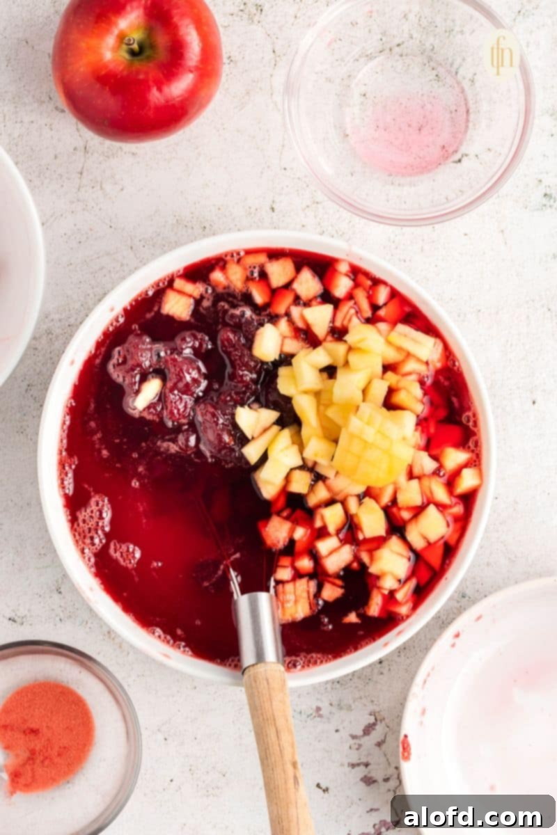 Raw ingredients of jello with fruit in a white bowl, showing cranberry sauce and chopped apples added to the gelatin mixture.