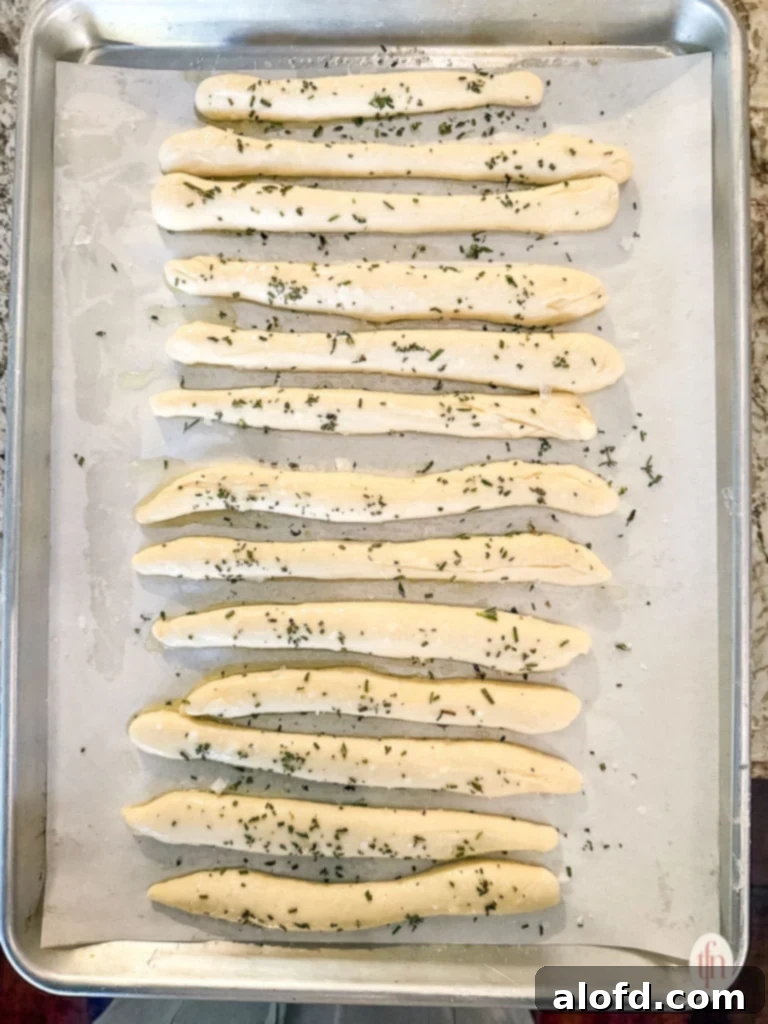 Effortless Charcuterie Boards 7 Uncooked breadstick dough pieces, seasoned with green herbs, arranged on a baking sheet lined with parchment paper.