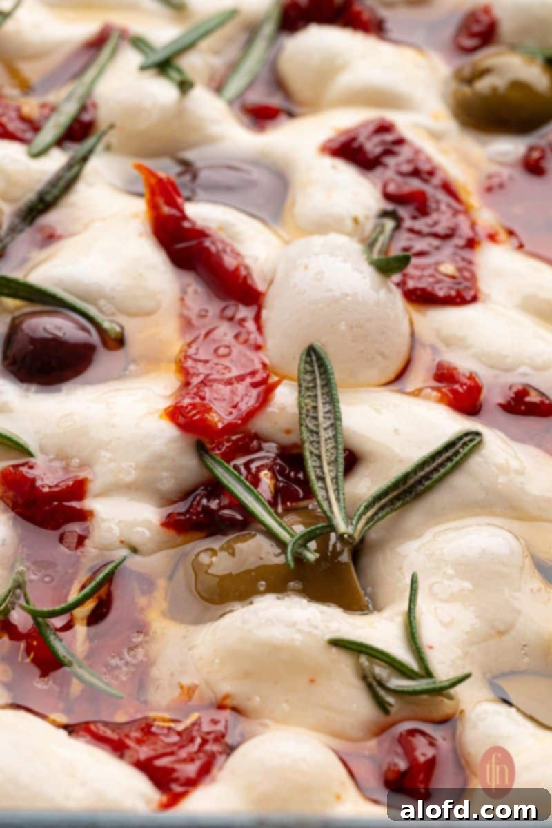 Close-up of unbaked sourdough focaccia dough, dimpled and topped with ingredients.