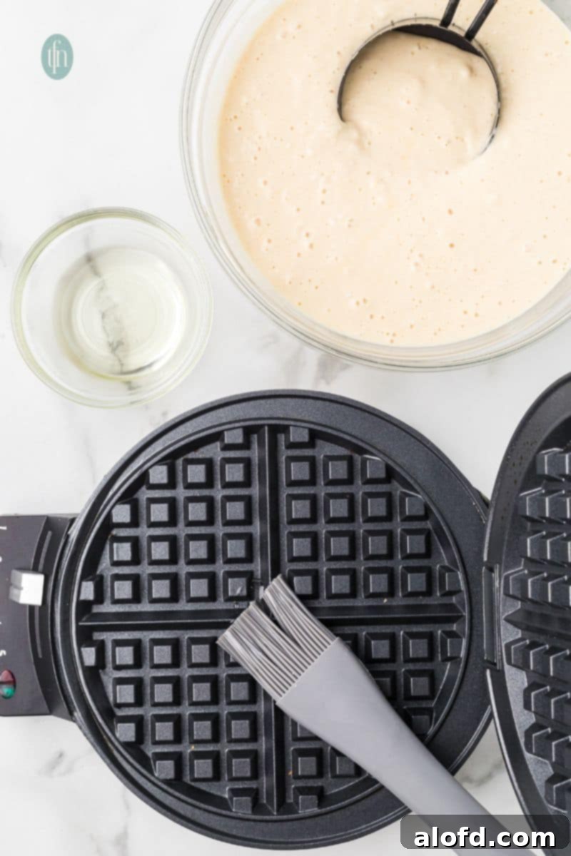 Fluffy Golden Bisquick Waffles 10 A waffle maker with a pastry brush and a bowl of batter mixture.
