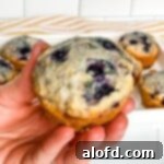 A square photograph of a hand gently holding a warm Bisquick blueberry muffin, with a tempting array of more muffins visible in the background.