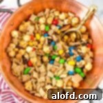 A close-up shot of a rustic copper bowl, complete with a handle, brimming with Chex Mix featuring M&Ms, accompanied by a golden scoop and a striped kitchen towel in the soft background.