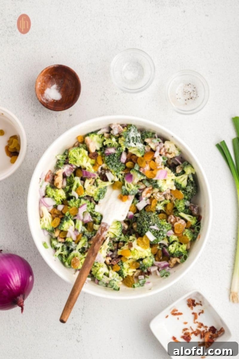 Tossing broccoli salad ingredients with a rubber spatula in a white mixing bowl, ensuring even coating of the dressing.