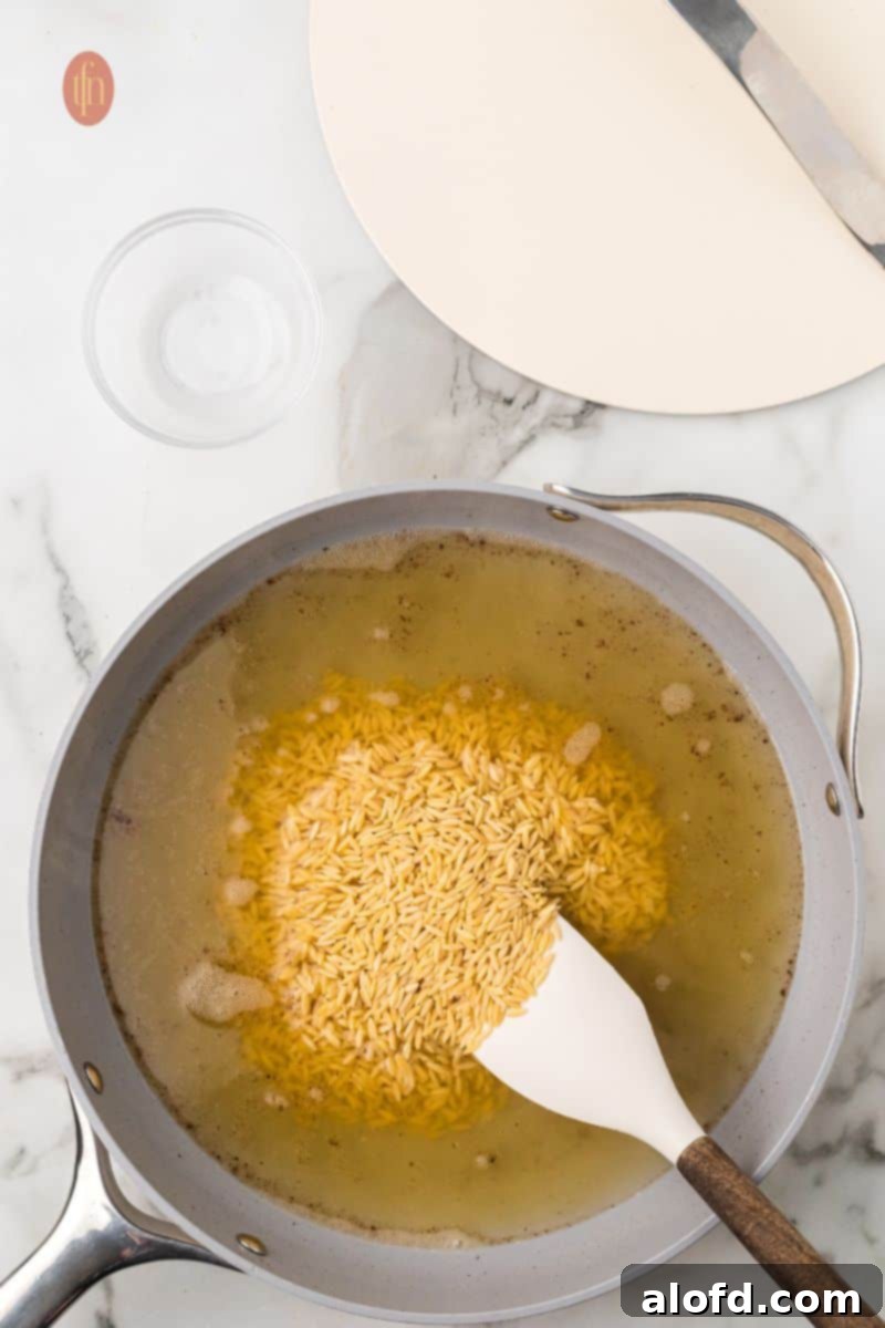 Zesty Lemon Chicken Orzo Pasta 15 Orzo pasta cooking in a pan with chicken broth, visually showing the absorption of liquid and the pasta beginning to soften.