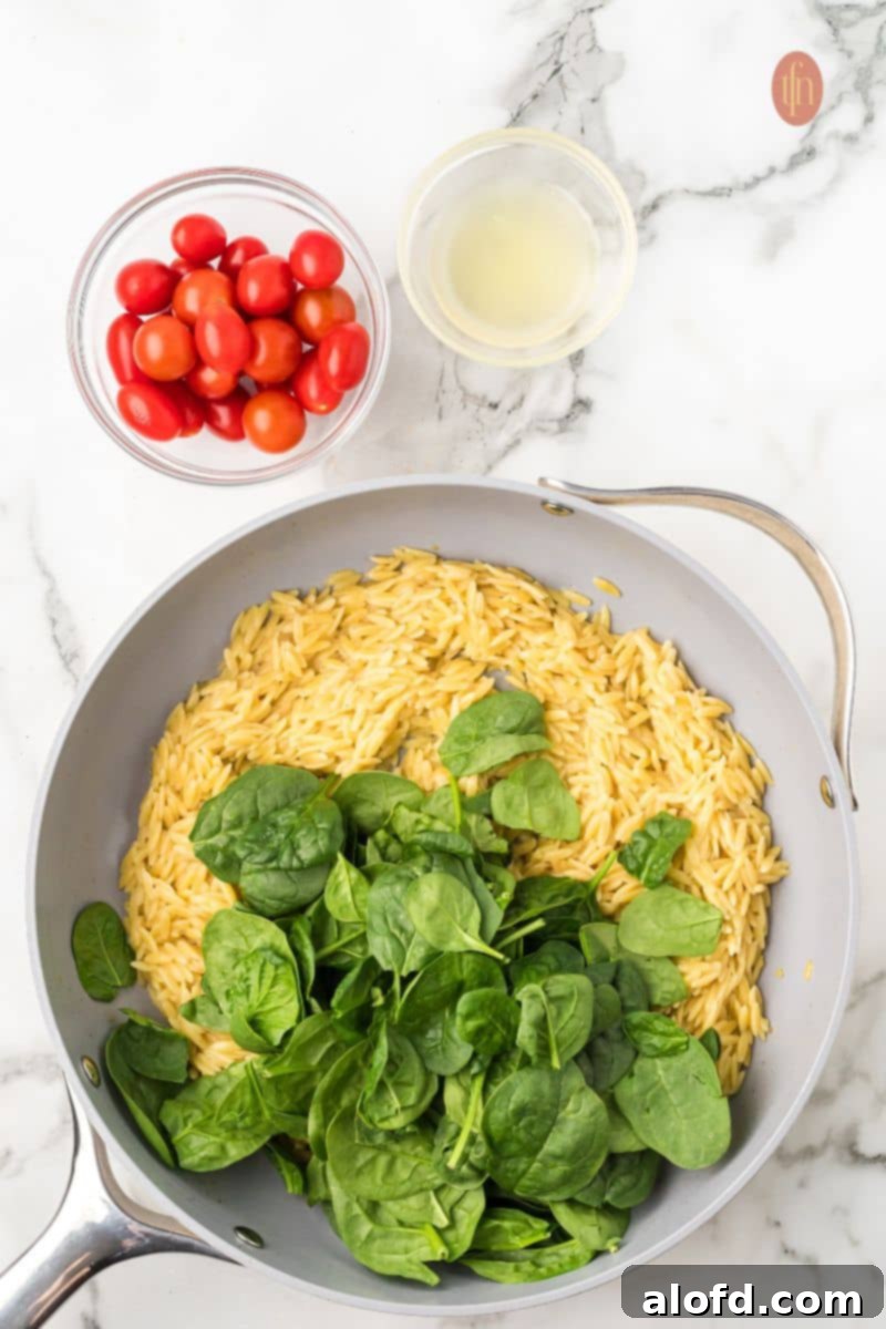 Zesty Lemon Chicken Orzo Pasta 17 Orzo pasta in a pan with baby spinach, next to bowls of liquid ingredients and fresh tomatoes, demonstrating the process of adding final fresh components.