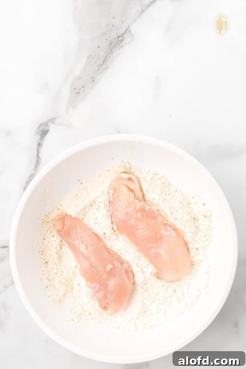 Zesty Lemon Chicken Orzo Pasta 9 Two pieces of raw chicken tenderloin being carefully coated in a bowl of seasoned flour mixture, ensuring even coverage.