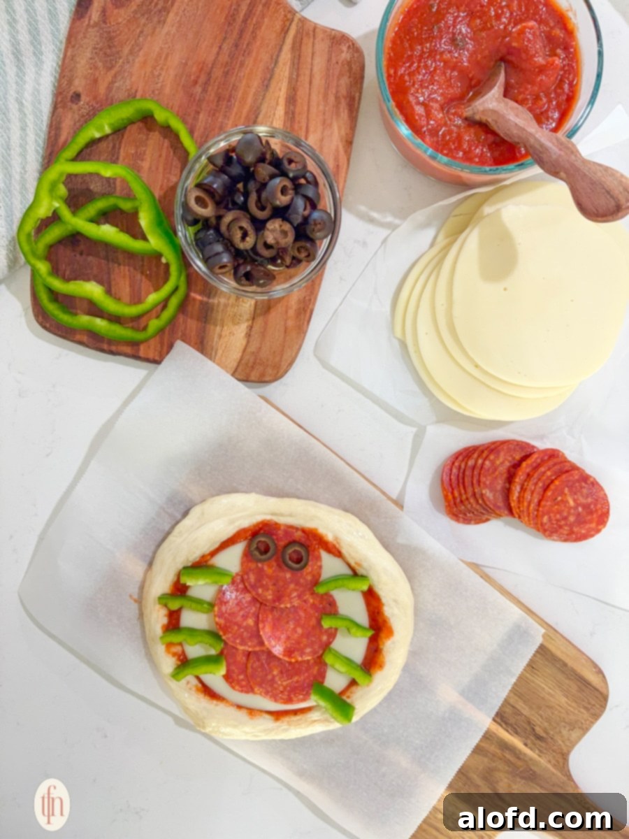 Monster Mash Pizza 11 An unbaked personal pizza is being prepared as a spider. The round dough has sauce and cheese, and the toppings are arranged to look like a spider with pepperoni for the body, olive slices for eyes, and green bell pepper strips for legs.