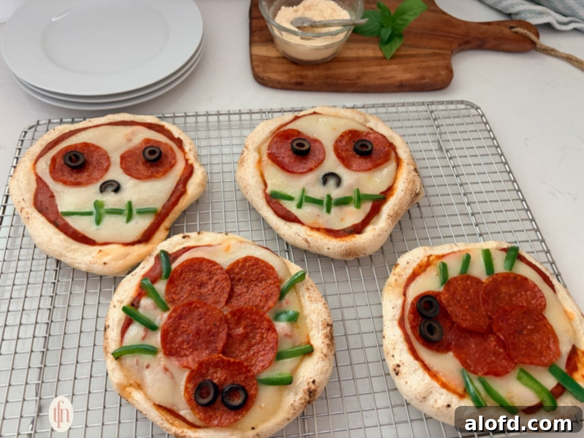 Monster Mash Pizza 12 A wide, eye-level shot of four Halloween-themed personal pizzas on a wire cooling rack. Two are skulls and two are spiders, all made with pepperoni, olives, and green peppers.